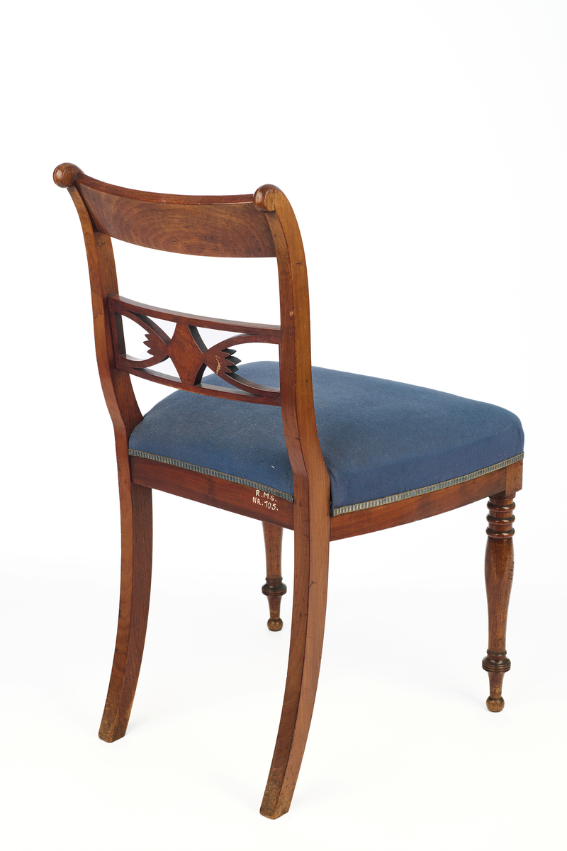 Stol av finért mahogni, som hviler på fire ben. Åpen rygg med glatt buet toppstykke med intarsia i lyst tre. Gjennombrutt tverrspross med halvsirkelformede ornamenter, hvor det ovale midtpartiet er markert med en riflet metallrosett. Kurvede akterstaver glir over i bakbenene, mens forbenene er dreide. Trapesformet sete med sekundært blåfarget trekk i ull. Mykt buet sarg, hvis sider er svakt trukket inn under setet.