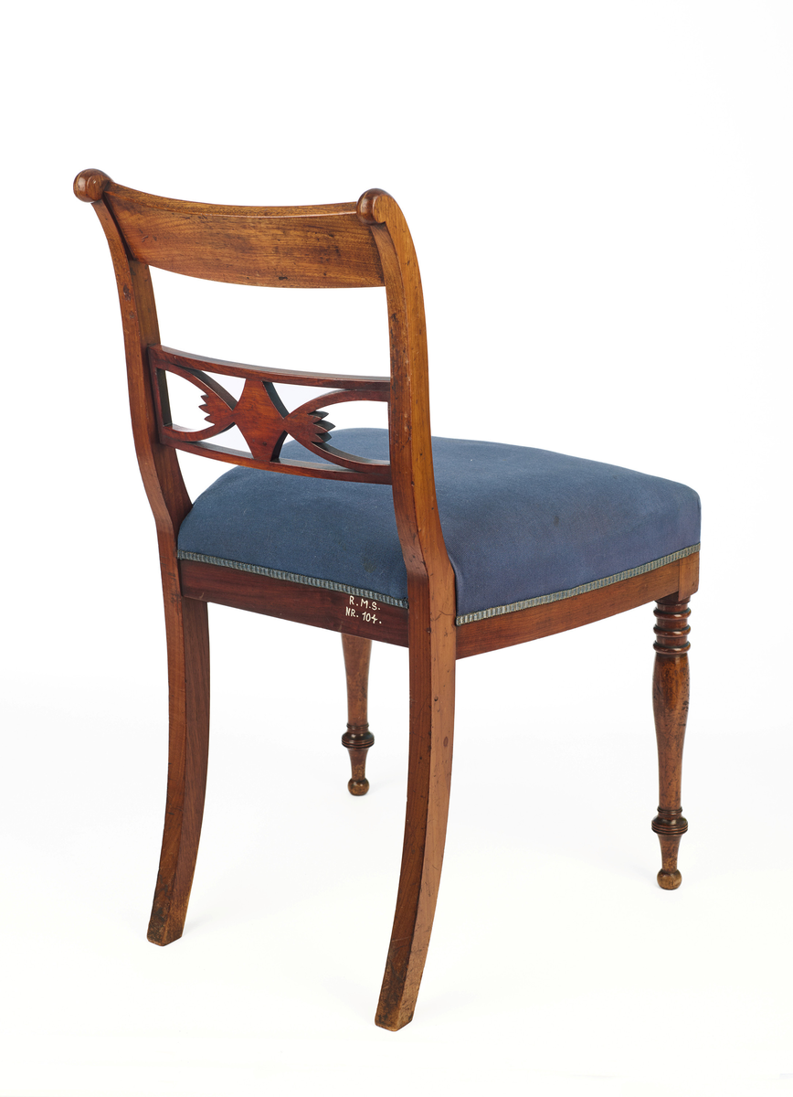 Stol av finért mahogni, som hviler på fire ben. Åpen rygg med glatt buet toppstykke med intarsia i lyst tre. Gjennombrutt tverrspross med halvsirkelformede ornamenter, hvor det ovale midtpartiet er markert med en riflet metallrosett. Kurvede akterstaver glir over i bakbenene, mens forbenene er dreide. Trapesformet sete med sekundært blåfarget trekk i ull. Mykt buet sarg, hvis sider er svakt trukket inn under setet.