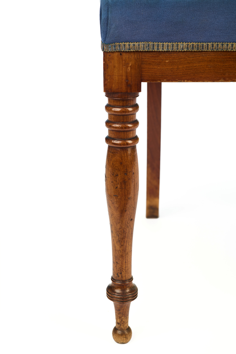 Stol av finért mahogni, som hviler på fire ben. Åpen rygg med glatt buet toppstykke med intarsia i lyst tre. Gjennombrutt tverrspross med halvsirkelformede ornamenter, hvor det ovale midtpartiet er markert med en riflet metallrosett. Kurvede akterstaver glir over i bakbenene, mens forbenene er dreide. Trapesformet sete med sekundært blåfarget trekk i ull. Mykt buet sarg, hvis sider er svakt trukket inn under setet.