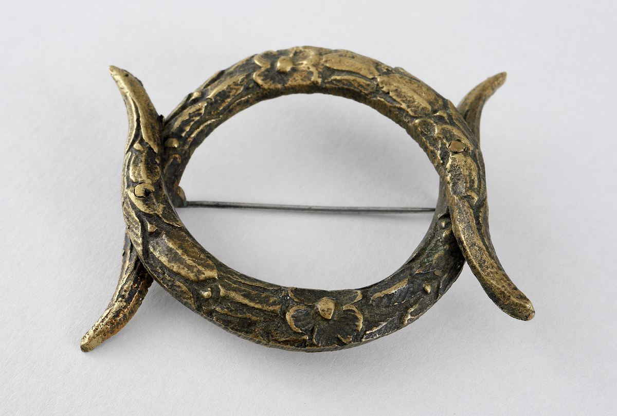 Spänne/brosch av gulmetall. Består av två stycken c-formade fastoner (bondflätor) nitade fast i varandra. Det är tillverkat av gulmetall och har en gångjärnsförsedd nål baktill som låses med en hake.

Inskrivet i huvudbok mellan 1898.