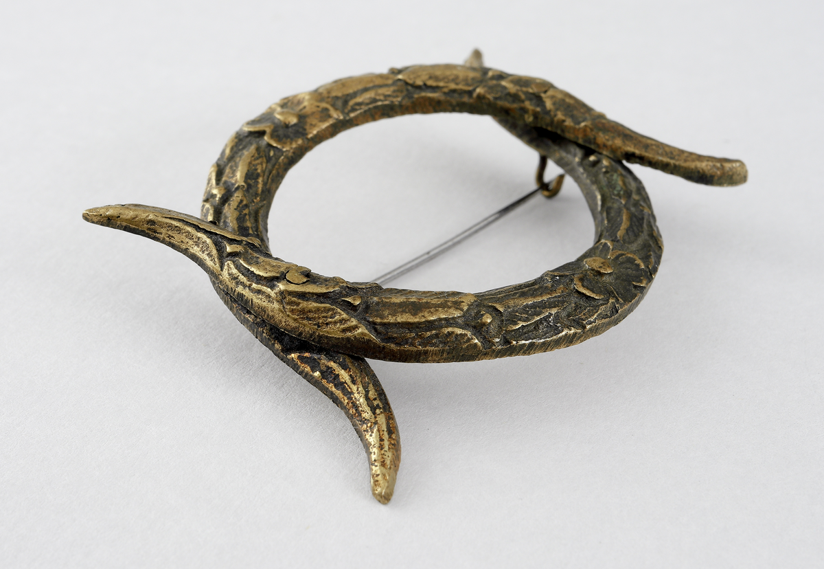 Spänne/brosch av gulmetall. Består av två stycken c-formade fastoner (bondflätor) nitade fast i varandra. Det är tillverkat av gulmetall och har en gångjärnsförsedd nål baktill som låses med en hake.

Inskrivet i huvudbok mellan 1898.