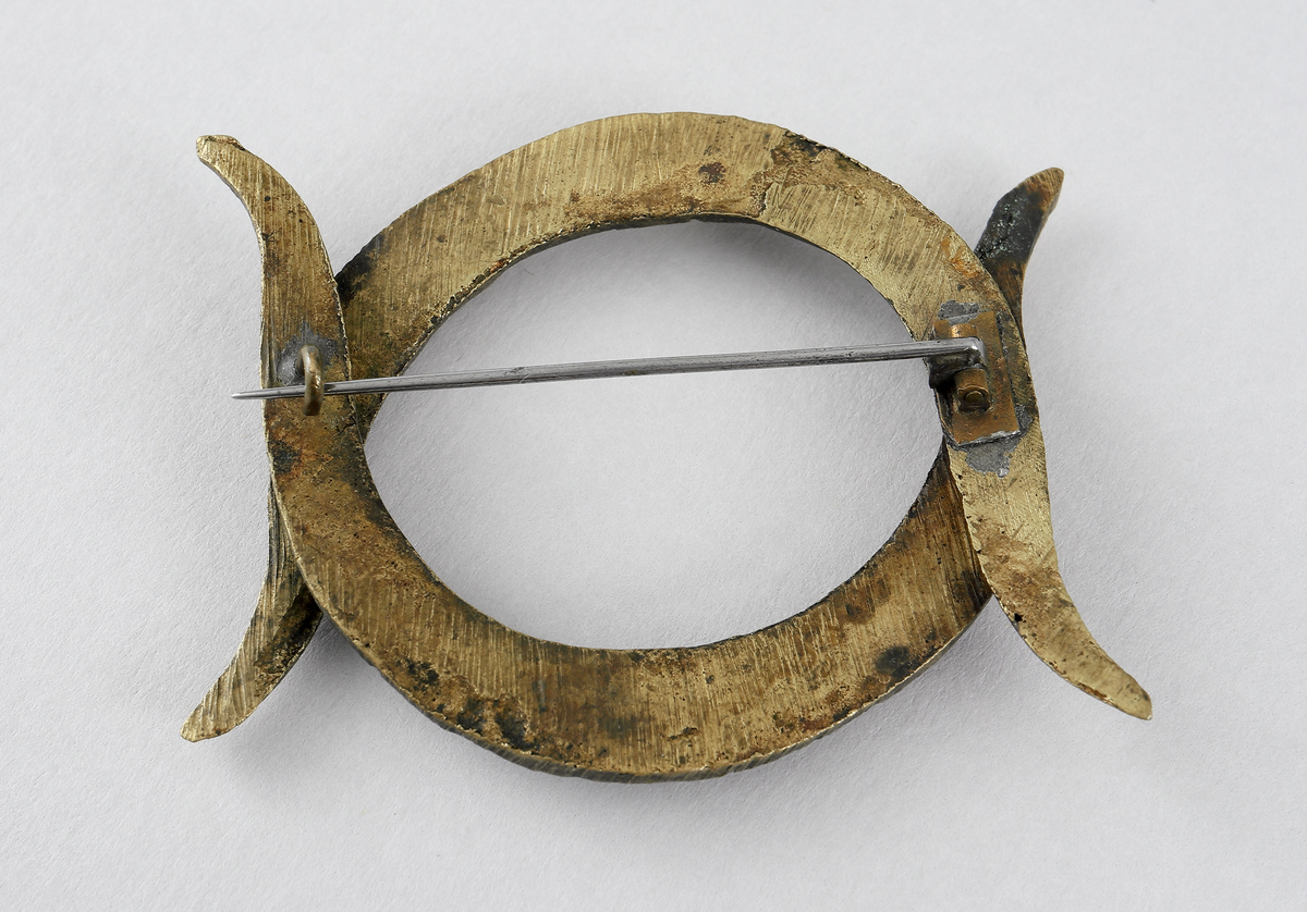 Spänne/brosch av gulmetall. Består av två stycken c-formade fastoner (bondflätor) nitade fast i varandra. Det är tillverkat av gulmetall och har en gångjärnsförsedd nål baktill som låses med en hake.

Inskrivet i huvudbok mellan 1898.