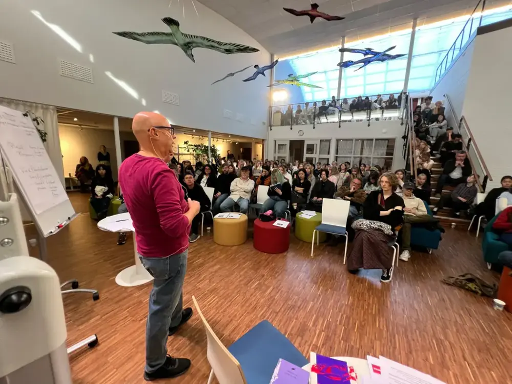 En mann i lilla genser snakker mot en gruppe ungdommer som sitter i salen og på en trapp. Et kunstverk med fugler henger fra taket. 