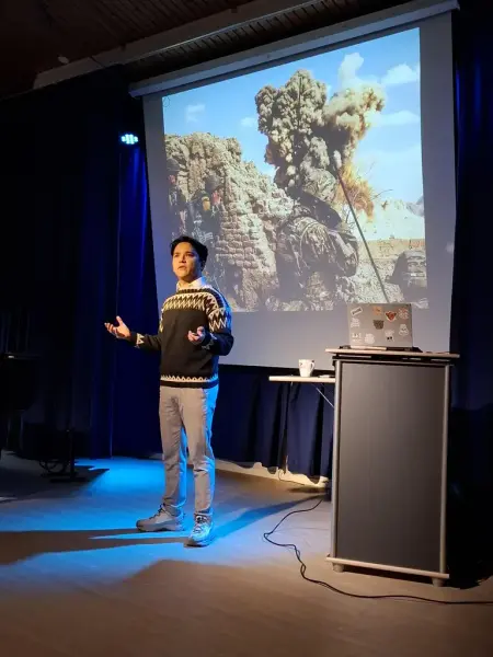 En ung mann står foran en stor skjerm med et krigsbilde på.