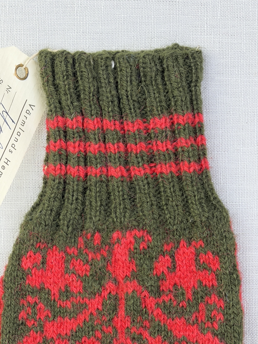 En stickad fingervante, med randig mudd i 2-2 resår (2 räta och 2 aviga maskor) och mönsterstickad vante i slätstickning, av 2-trådigt ullgarn.