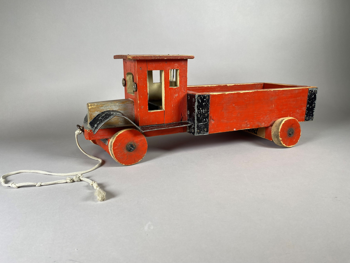 Lastebil snekret i tre med metallbeslag. Treverket er rødmalt, med svarte beslag. Lekebilen har to nummerskilt foran og bak med merking "U54227". På undersiden er gjenstanden merket med et påskrevet navn. På ene siden av lasteplanet er det festet et merke med Postverkets logomerke. Lekebilen har noe slitasjemerker, trolig etter bruk.