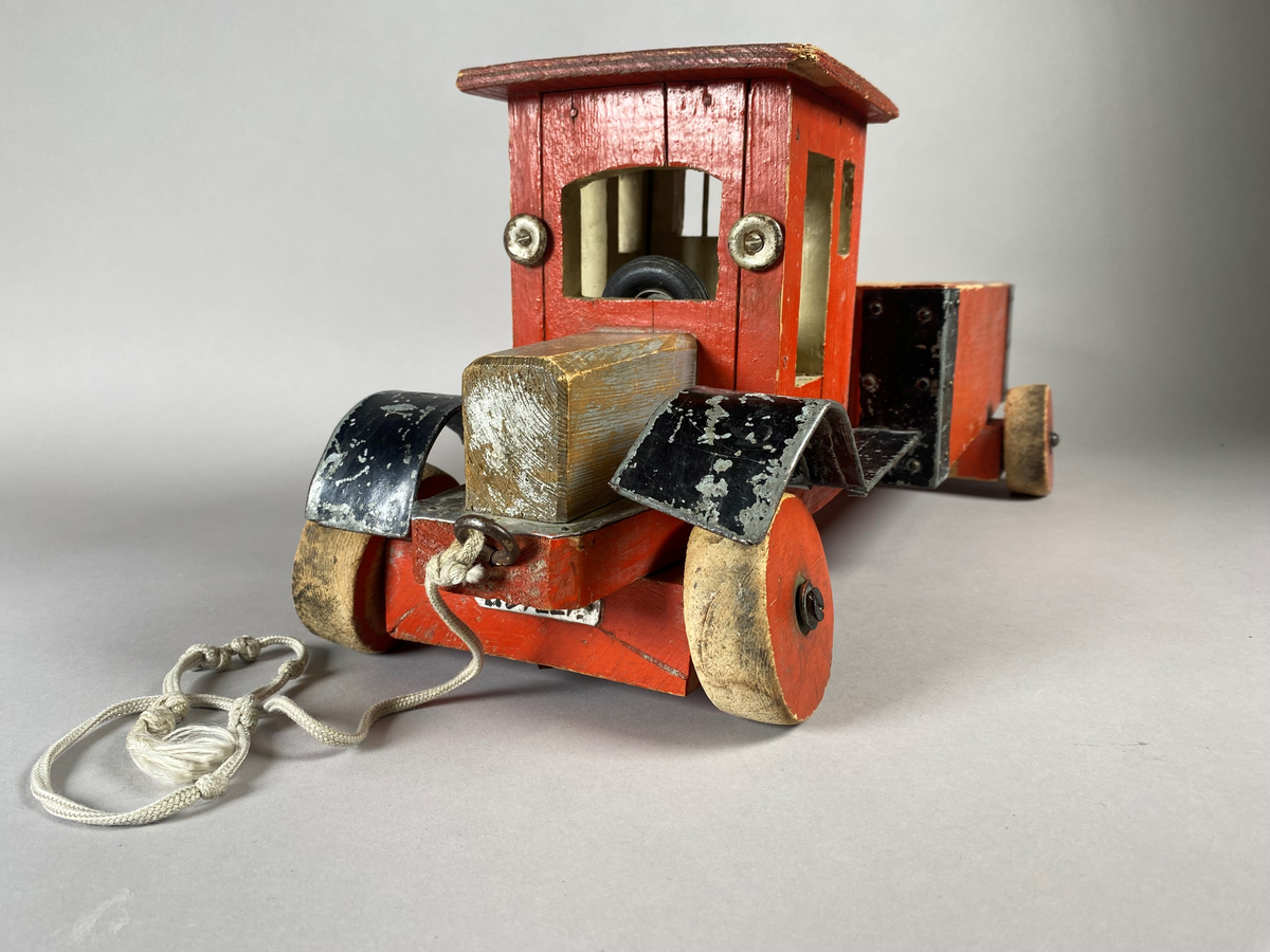 Lastebil snekret i tre med metallbeslag. Treverket er rødmalt, med svarte beslag. Lekebilen har to nummerskilt foran og bak med merking "U54227". På undersiden er gjenstanden merket med et påskrevet navn. På ene siden av lasteplanet er det festet et merke med Postverkets logomerke. Lekebilen har noe slitasjemerker, trolig etter bruk.