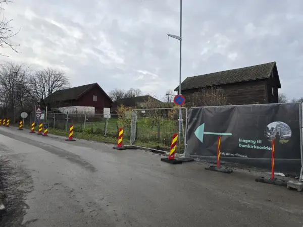 Fra parkeringsplassen i Strandvegen 100 er det et stort banner på et byggegjerde med pil som peker til venstre nedover Strandvegen mot Mjøsa. Langs høyre vegkant står det sperringer som er ment for å trygge fotgjengere ved at de kan gå på innsiden av sperringene.