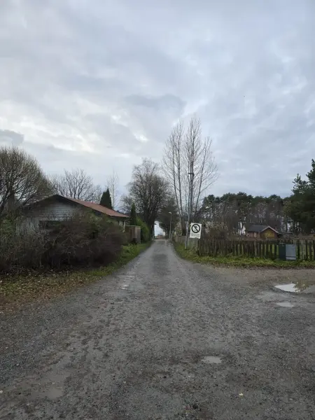 Grusveg ned mot Mjøsa mellom en hekk og garasje, og et gjerde. Skilt med "Innkjøring forbudt uten avtale med museet".