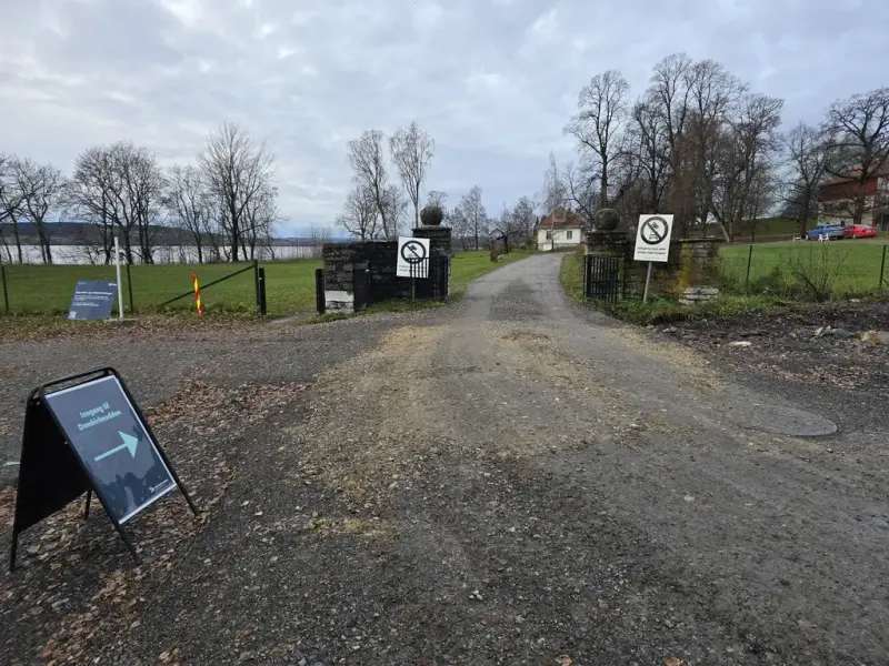 Grusveg som fører fra Strandvegen og inn på museumsområdet gjennom en port. Plakat med pil og teksten "inngang til Domkirkeodden" vises til venstre i bildet, rett fram fra porten skimter man Storhamarstuene, og oppe til høyre Aulaen på Domkirkeodden.
