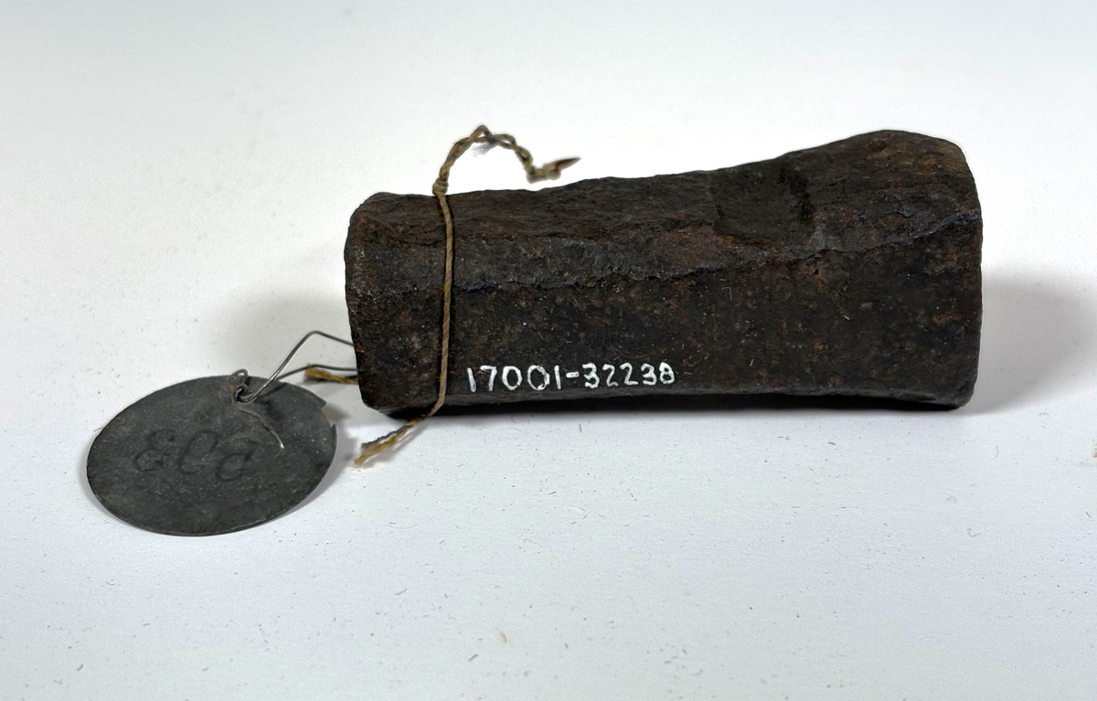 Rektangulärt metallstycke med en cirkelformad slagyta i ena änden. Den andra änden är något bredare och har en instansad text "SB" krönt med en kungakrona.