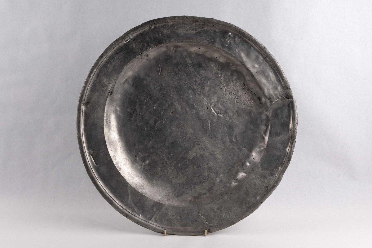Stort, sirkulært serveringsbrett funnet ved skipet AKERENDAM som forliste på Runde i 1725. Tinnfat med flat bunn og 5 cm. bred rand. To stempler på undersiden. Motiv er en engel med vinger, holder noe i hendene. Det andre stempelet er blomst og krone. Brett med bulker/skader.