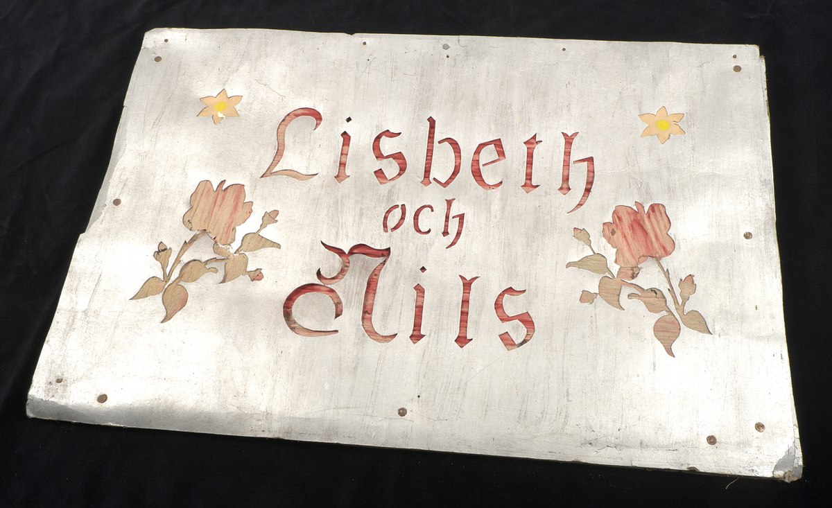 Bröllopslykta/transparang av kraftigt silverfärgat papper spänt över träram. I pappret finns skurna dekorationer i form av florala mönster, symboler och text. Bakom de utskurna partierna har silkespapper i grönt, rött och gult monterats. På framsidan återfinns texten: "Lisbeth och Nils", på baksidan: "Lycka och wälgång".