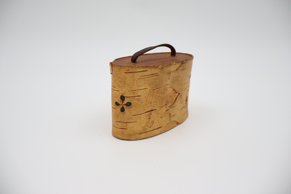 Oval näverburk med lock. Liten. Nävret är tampat stående. Nävret är pålimmat på bottenplattan i trä. Även locket består av trä och har ett handtag i läder. Läderremmen är trädd genom två hål i locket och på lockets insida avslutas remmen med två knutar. Burken har utstansade dekorationer i form av solfjädrar, romber, blommor och en stjärna. Burken består av två lager näver och emellan nävret är blått tyg inlagt som skymtar fram i stjärnan och blommorna. Burken är i relativt gott skick.