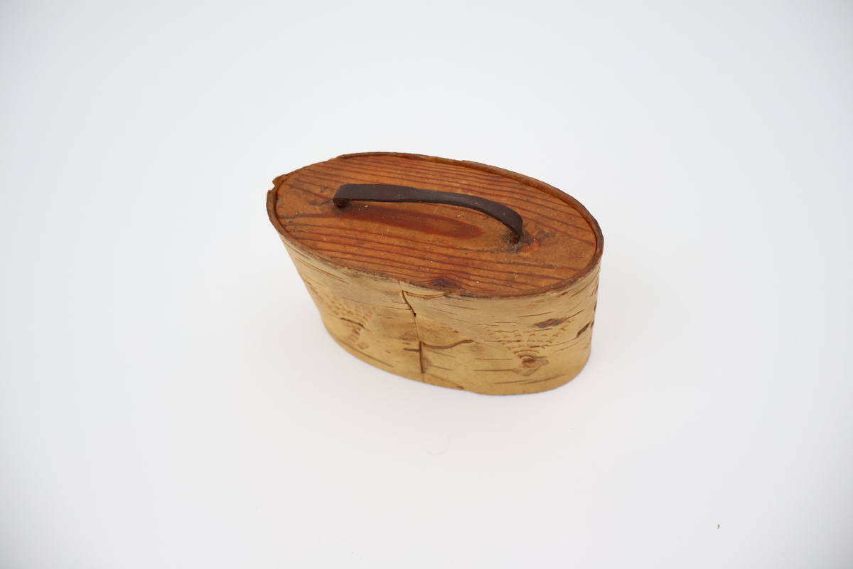 Oval näverburk med lock. Liten. Nävret är tampat stående. Nävret är pålimmat på bottenplattan i trä. Även locket består av trä och har ett handtag i läder. Läderremmen är trädd genom två hål i locket och på lockets insida avslutas remmen med två knutar. Burken har utstansade dekorationer i form av solfjädrar, romber, blommor och en stjärna. Burken består av två lager näver och emellan nävret är blått tyg inlagt som skymtar fram i stjärnan och blommorna. Burken är i relativt gott skick.