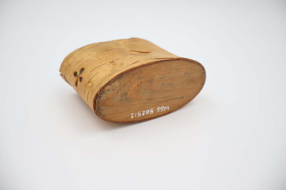 Oval näverburk med lock. Liten. Nävret är tampat stående. Nävret är pålimmat på bottenplattan i trä. Även locket består av trä och har ett handtag i läder. Läderremmen är trädd genom två hål i locket och på lockets insida avslutas remmen med två knutar. Burken har utstansade dekorationer i form av solfjädrar, romber, blommor och en stjärna. Burken består av två lager näver och emellan nävret är blått tyg inlagt som skymtar fram i stjärnan och blommorna. Burken är i relativt gott skick.