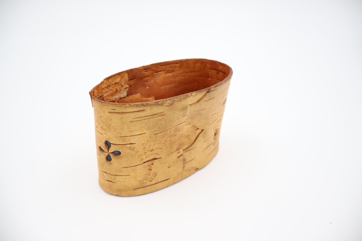 Oval näverburk med lock. Liten. Nävret är tampat stående. Nävret är pålimmat på bottenplattan i trä. Även locket består av trä och har ett handtag i läder. Läderremmen är trädd genom två hål i locket och på lockets insida avslutas remmen med två knutar. Burken har utstansade dekorationer i form av solfjädrar, romber, blommor och en stjärna. Burken består av två lager näver och emellan nävret är blått tyg inlagt som skymtar fram i stjärnan och blommorna. Burken är i relativt gott skick.