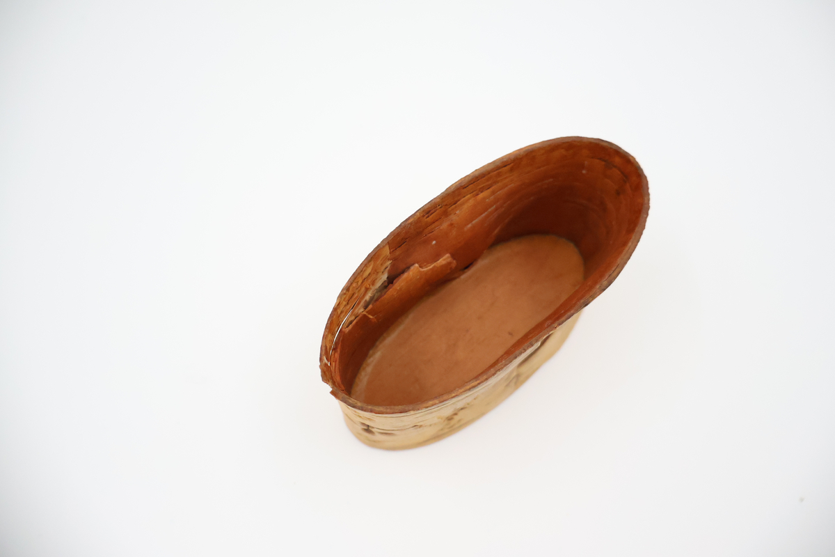 Oval näverburk med lock. Liten. Nävret är tampat stående. Nävret är pålimmat på bottenplattan i trä. Även locket består av trä och har ett handtag i läder. Läderremmen är trädd genom två hål i locket och på lockets insida avslutas remmen med två knutar. Burken har utstansade dekorationer i form av solfjädrar, romber, blommor och en stjärna. Burken består av två lager näver och emellan nävret är blått tyg inlagt som skymtar fram i stjärnan och blommorna. Burken är i relativt gott skick.