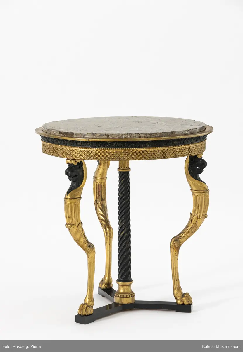 KLM 12977. Bord. Runt bord med stomme av furu. Skurna skulpterade delar troligen av lövträ. Sarg och ben rikt skulpterade och helförgyllda. Svärtade delar målade med svart oljefärg direkt på stommen. Ytskikt består av pastellage på bordssarg. Grundering med limkrita under polimentförgyllda delar, bladguld. 

Bordet bärs till synes av tre tunna smäckra getbockben, som avslutas med en lejontass. Undertill stabiliserat av en spiralkannelerad, lätt avsmalnade upptill svärtad matt kolonn, stående på ett förgyllt doriskt kapitäl, med tillagd pärllist och en förgylld kannelerad kolonnbas under bordskivan, dvs. ett upp och nedvänt motiv. Stödet bär upp den tunga stenskivan. Detta fjärde bordsben är skymt av de övriga tre benen vid rakt betraktande framifrån. De fyra benen är förankrade mot golvet på en svartmålat högblankpolerat rakt avslutat trepass av trä, som reflekterar de förgyllda benen. Trepasset vilar i sin tur på låga slitklossar, en under varje lejontass och under bordets mitt.

Lejontassen övergår i ett bockben, vid knäleden övergår formen till en hornformation som utgörs av släta aningen veckade långsträckta blad, ur denna bladform utgår en kannelerad kolonn som avslutas med en abakus, ur denna, i sin tur, växer ett svärtat lejonhuvud iförd ett egyptiskt farao-dok, doket rullas samman baktill till ett joniskt kapitäl som vilar på lejonets huvud, vilket i sin tur bär upp bordets sarg. De tre bockbenen är polimentförgyllda i sin helhet förutom lejonhuvudet som är mattsvärtat. Sargen som tar vid efter de joniska kapitälen, har i närmsta anslutning, en bred, rak, förgylld pastellagelist med upprepningsmönster av stiliserade granatäppelkärnor. Denna del avslutas upptill med en förgylld pärllist. Över den förgyllda pärllisten kragar en svärtad list med stående akantus, lätt konvex utförd i pastellage. Denna bär i sin tur en cirkelformad utskjutande förgylld platt kraglist, på vilken den något indragna och lätt förhöjda stenskivan vilar. Märkning och hallstämpel saknas.

Stenskiva av polerad revkalksten, Gotland, Slite, ”Megalomuskalksten”. Skal av fossiliserade musslor och ostron är synliga. Undersida skråhuggen. Bordsskivan är utförd i stucco lustro, en puts- och målningsteknik som ger en glänsande yta och imiterar marmor.