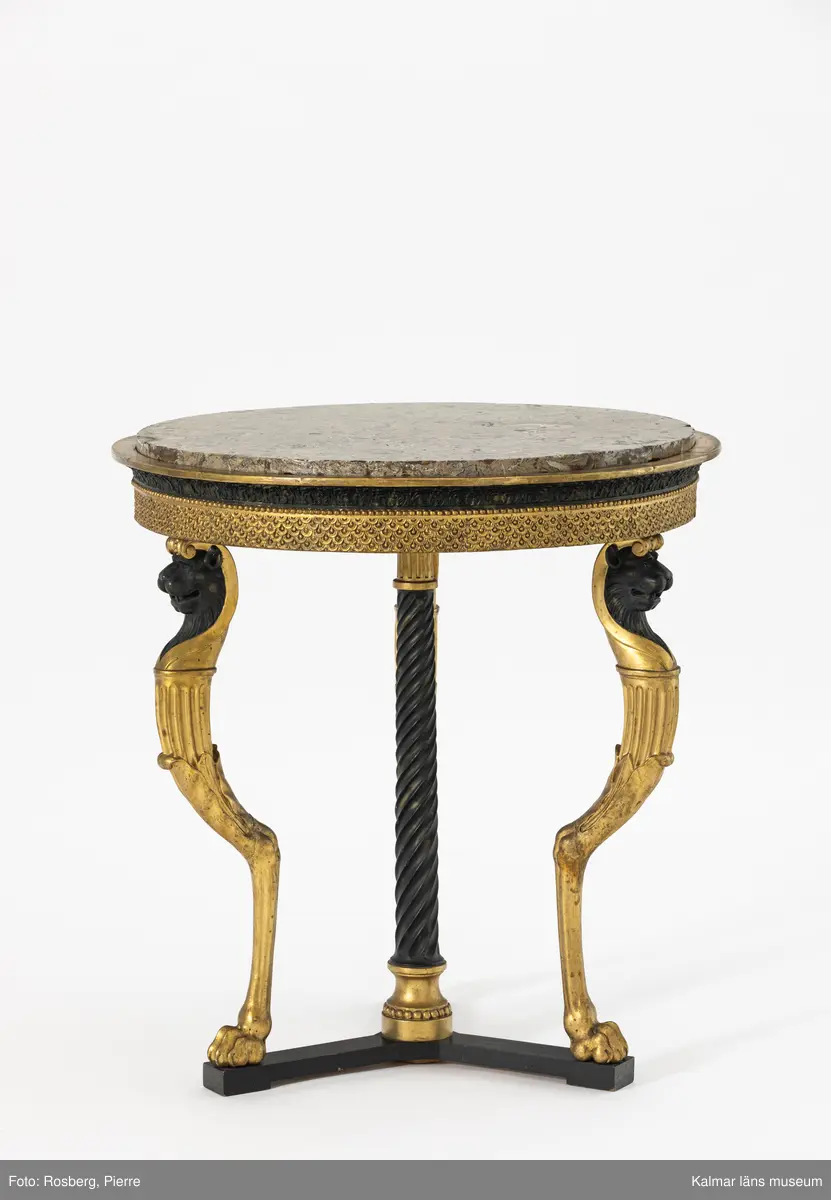 KLM 12977. Bord. Runt bord med stomme av furu. Skurna skulpterade delar troligen av lövträ. Sarg och ben rikt skulpterade och helförgyllda. Svärtade delar målade med svart oljefärg direkt på stommen. Ytskikt består av pastellage på bordssarg. Grundering med limkrita under polimentförgyllda delar, bladguld. 

Bordet bärs till synes av tre tunna smäckra getbockben, som avslutas med en lejontass. Undertill stabiliserat av en spiralkannelerad, lätt avsmalnade upptill svärtad matt kolonn, stående på ett förgyllt doriskt kapitäl, med tillagd pärllist och en förgylld kannelerad kolonnbas under bordskivan, dvs. ett upp och nedvänt motiv. Stödet bär upp den tunga stenskivan. Detta fjärde bordsben är skymt av de övriga tre benen vid rakt betraktande framifrån. De fyra benen är förankrade mot golvet på en svartmålat högblankpolerat rakt avslutat trepass av trä, som reflekterar de förgyllda benen. Trepasset vilar i sin tur på låga slitklossar, en under varje lejontass och under bordets mitt.

Lejontassen övergår i ett bockben, vid knäleden övergår formen till en hornformation som utgörs av släta aningen veckade långsträckta blad, ur denna bladform utgår en kannelerad kolonn som avslutas med en abakus, ur denna, i sin tur, växer ett svärtat lejonhuvud iförd ett egyptiskt farao-dok, doket rullas samman baktill till ett joniskt kapitäl som vilar på lejonets huvud, vilket i sin tur bär upp bordets sarg. De tre bockbenen är polimentförgyllda i sin helhet förutom lejonhuvudet som är mattsvärtat. Sargen som tar vid efter de joniska kapitälen, har i närmsta anslutning, en bred, rak, förgylld pastellagelist med upprepningsmönster av stiliserade granatäppelkärnor. Denna del avslutas upptill med en förgylld pärllist. Över den förgyllda pärllisten kragar en svärtad list med stående akantus, lätt konvex utförd i pastellage. Denna bär i sin tur en cirkelformad utskjutande förgylld platt kraglist, på vilken den något indragna och lätt förhöjda stenskivan vilar. Märkning och hallstämpel saknas.

Stenskiva av polerad revkalksten, Gotland, Slite, ”Megalomuskalksten”. Skal av fossiliserade musslor och ostron är synliga. Undersida skråhuggen. Bordsskivan är utförd i stucco lustro, en puts- och målningsteknik som ger en glänsande yta och imiterar marmor.