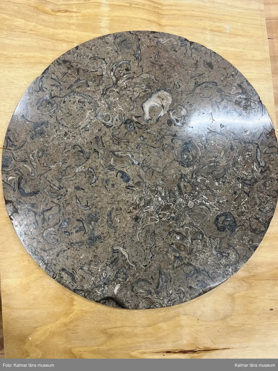 KLM 12977. Bord. Runt bord med stomme av furu. Skurna skulpterade delar troligen av lövträ. Sarg och ben rikt skulpterade och helförgyllda. Svärtade delar målade med svart oljefärg direkt på stommen. Ytskikt består av pastellage på bordssarg. Grundering med limkrita under polimentförgyllda delar, bladguld. 

Bordet bärs till synes av tre tunna smäckra getbockben, som avslutas med en lejontass. Undertill stabiliserat av en spiralkannelerad, lätt avsmalnade upptill svärtad matt kolonn, stående på ett förgyllt doriskt kapitäl, med tillagd pärllist och en förgylld kannelerad kolonnbas under bordskivan, dvs. ett upp och nedvänt motiv. Stödet bär upp den tunga stenskivan. Detta fjärde bordsben är skymt av de övriga tre benen vid rakt betraktande framifrån. De fyra benen är förankrade mot golvet på en svartmålat högblankpolerat rakt avslutat trepass av trä, som reflekterar de förgyllda benen. Trepasset vilar i sin tur på låga slitklossar, en under varje lejontass och under bordets mitt.

Lejontassen övergår i ett bockben, vid knäleden övergår formen till en hornformation som utgörs av släta aningen veckade långsträckta blad, ur denna bladform utgår en kannelerad kolonn som avslutas med en abakus, ur denna, i sin tur, växer ett svärtat lejonhuvud iförd ett egyptiskt farao-dok, doket rullas samman baktill till ett joniskt kapitäl som vilar på lejonets huvud, vilket i sin tur bär upp bordets sarg. De tre bockbenen är polimentförgyllda i sin helhet förutom lejonhuvudet som är mattsvärtat. Sargen som tar vid efter de joniska kapitälen, har i närmsta anslutning, en bred, rak, förgylld pastellagelist med upprepningsmönster av stiliserade granatäppelkärnor. Denna del avslutas upptill med en förgylld pärllist. Över den förgyllda pärllisten kragar en svärtad list med stående akantus, lätt konvex utförd i pastellage. Denna bär i sin tur en cirkelformad utskjutande förgylld platt kraglist, på vilken den något indragna och lätt förhöjda stenskivan vilar. Märkning och hallstämpel saknas.

Stenskiva av polerad revkalksten, Gotland, Slite, ”Megalomuskalksten”. Skal av fossiliserade musslor och ostron är synliga. Undersida skråhuggen. Bordsskivan är utförd i stucco lustro, en puts- och målningsteknik som ger en glänsande yta och imiterar marmor.