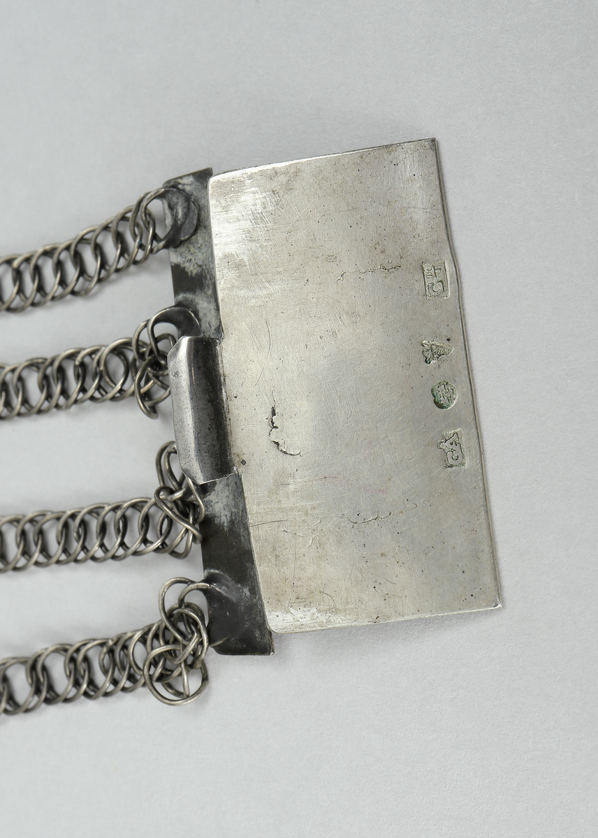 Halslås bestående av fyra länkar av olika längd, fästa på ett rektangulärt spänne av förgyllt silver. På spännet finns åtta upphöjda hjärtan omgivet av blomornament, runt kanten en bård av punktlinjer. I spännets nedre kant en kedja med ett hjärta i förgyllt silver.
Inskrivet i huvudkatalog 1938.