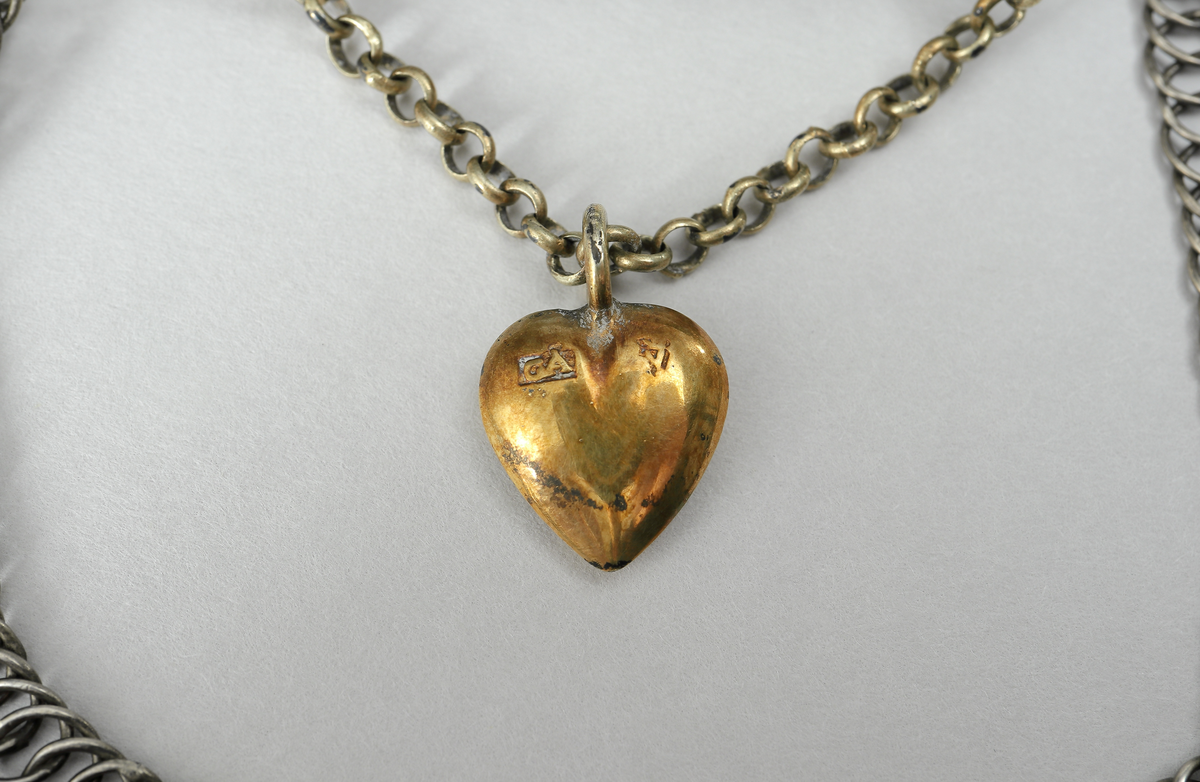 Halslås bestående av fyra länkar av olika längd, fästa på ett rektangulärt spänne av förgyllt silver. På spännet finns åtta upphöjda hjärtan omgivet av blomornament, runt kanten en bård av punktlinjer. I spännets nedre kant en kedja med ett hjärta i förgyllt silver.
Inskrivet i huvudkatalog 1938.