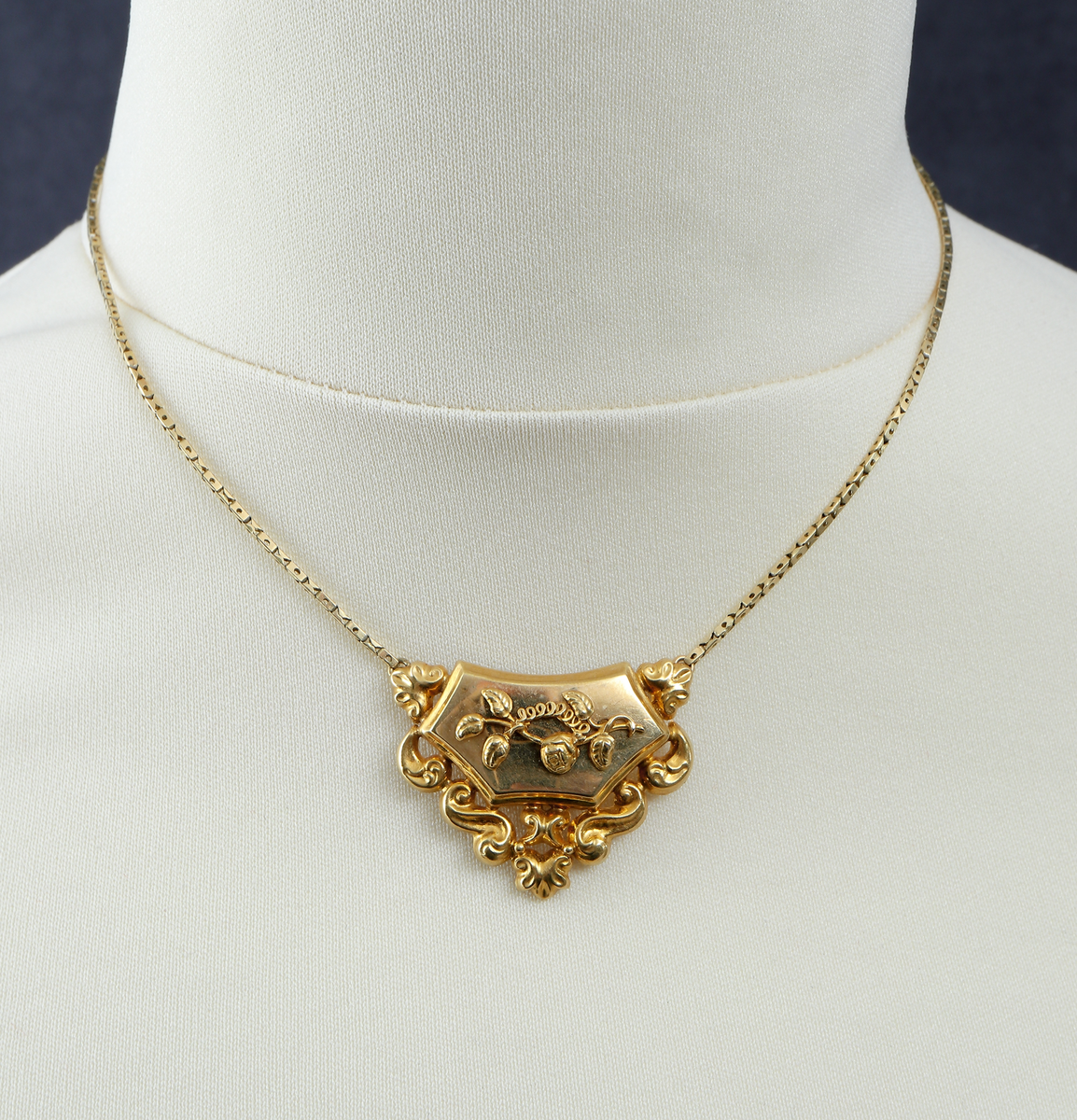 Halssmycke med kedja, tunnt guldbleck 18K, pressat. Bakpå skölden samt på ena låsdelen finns följande stämplar. Kontrollstämpel, 18 k, stadsstämpel Stockholm, Mästarstämpel EWZ, årtalsstämpel N4. Sköldformat med påritad dekor föreställande ros på gren. Runt sköljden s-formade utsmyckningar. Kedjan består av rektangulära genombrutna länkar. Låset är utformat som två mötande händer. 
Inskrivet i huvudkatalog 1963.