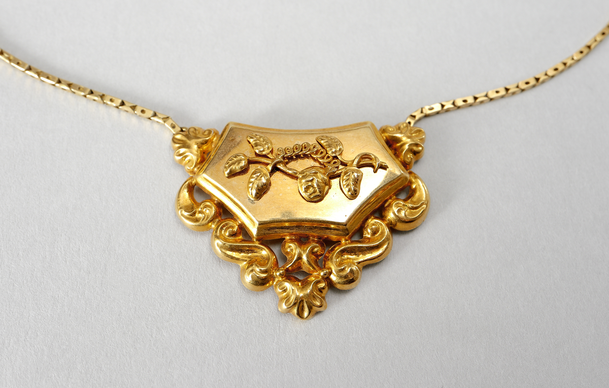 Halssmycke med kedja, tunnt guldbleck 18K, pressat. Bakpå skölden samt på ena låsdelen finns följande stämplar. Kontrollstämpel, 18 k, stadsstämpel Stockholm, Mästarstämpel EWZ, årtalsstämpel N4. Sköldformat med påritad dekor föreställande ros på gren. Runt sköljden s-formade utsmyckningar. Kedjan består av rektangulära genombrutna länkar. Låset är utformat som två mötande händer. 
Inskrivet i huvudkatalog 1963.