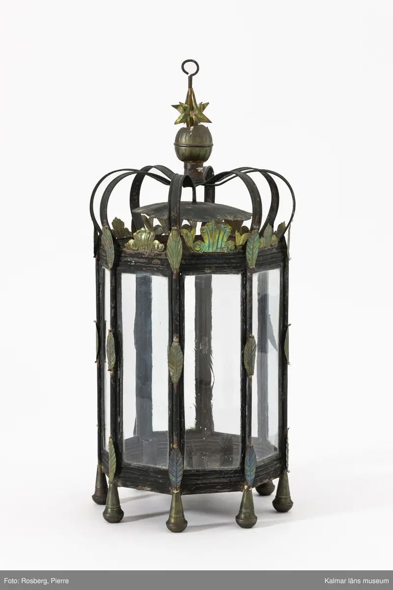 KLM 46902. Lykta. Åttakantig stomme av svartmålad metall med blyinfattade munblåsta rektangulära glasrutor, varav en försedd med gångjärn som dörr. Upptill bågformade band av grönmålad metall som möts i mitten som är försedd med en krok för upphängning. Lyktan är krönt med en kula och stjärnor av mässing. Runt om lyktan fastlödda mässingsdekorationer med pressat mönster i form av blad. Fötter av mässing. Inuti plats för ett större ljus samt tre mindre.