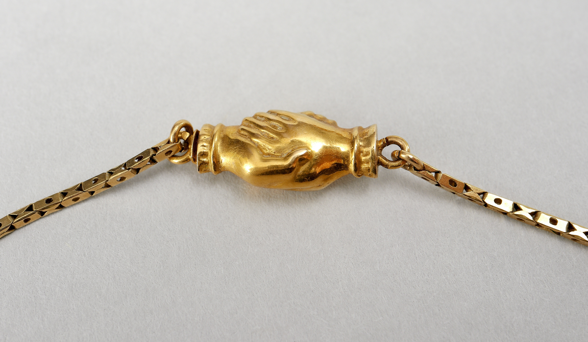 Halssmycke med kedja, tunnt guldbleck 18K, pressat. Bakpå skölden samt på ena låsdelen finns följande stämplar. Kontrollstämpel, 18 k, stadsstämpel Stockholm, Mästarstämpel EWZ, årtalsstämpel N4. Sköldformat med påritad dekor föreställande ros på gren. Runt sköljden s-formade utsmyckningar. Kedjan består av rektangulära genombrutna länkar. Låset är utformat som två mötande händer. 
Inskrivet i huvudkatalog 1963.