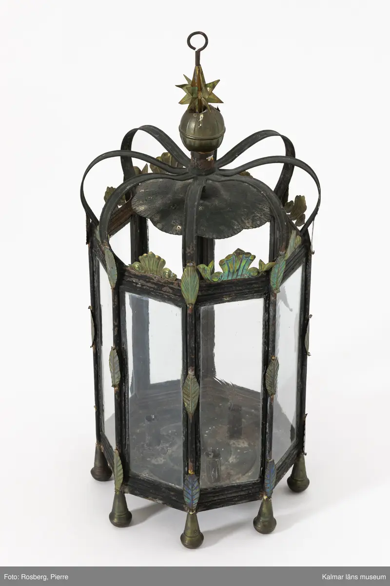 KLM 46902. Lykta. Åttakantig stomme av svartmålad metall med blyinfattade munblåsta rektangulära glasrutor, varav en försedd med gångjärn som dörr. Upptill bågformade band av grönmålad metall som möts i mitten som är försedd med en krok för upphängning. Lyktan är krönt med en kula och stjärnor av mässing. Runt om lyktan fastlödda mässingsdekorationer med pressat mönster i form av blad. Fötter av mässing. Inuti plats för ett större ljus samt tre mindre.