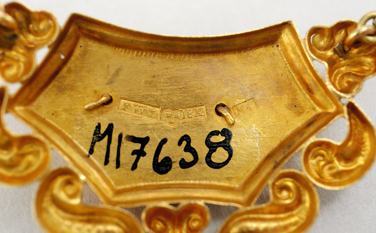 Halssmycke med kedja, tunnt guldbleck 18K, pressat. Bakpå skölden samt på ena låsdelen finns följande stämplar. Kontrollstämpel, 18 k, stadsstämpel Stockholm, Mästarstämpel EWZ, årtalsstämpel N4. Sköldformat med påritad dekor föreställande ros på gren. Runt sköljden s-formade utsmyckningar. Kedjan består av rektangulära genombrutna länkar. Låset är utformat som två mötande händer. 
Inskrivet i huvudkatalog 1963.