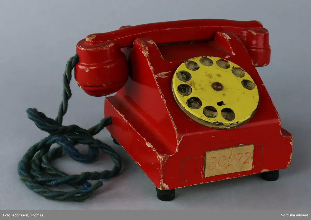 Leksakstelefon med nummerskiva och vidhängande telefonlur som sitter fast med en tvinnad lina av grönt garn. Etikett på framsidan med givarens telefonnummer textad med röd penna: "30272". På undersidan av telefonen finns en hålighet som ursprungligen kan ha innehållit en ringklocka. På undersidan är tre stöd av bakelit fastspikade, det fjärde saknas.
