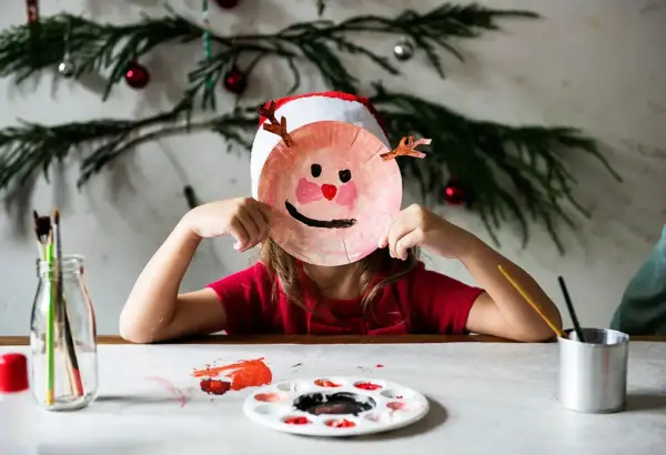 Barn som håller en målad papperstallrik framför ansiktet, med målarfärg och penslar på bordet.