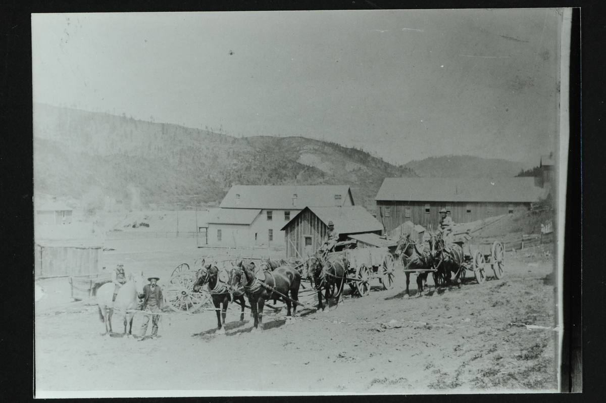 En gård med lys, to-etasjes hovedbygning, stor låve og flere uthus. I forgrunn to vogner med tilsammen fem hester foran. Til venstre en mann som leier en hest med et barn på. I bakgrunn en åsrygg med spredte trær. 
