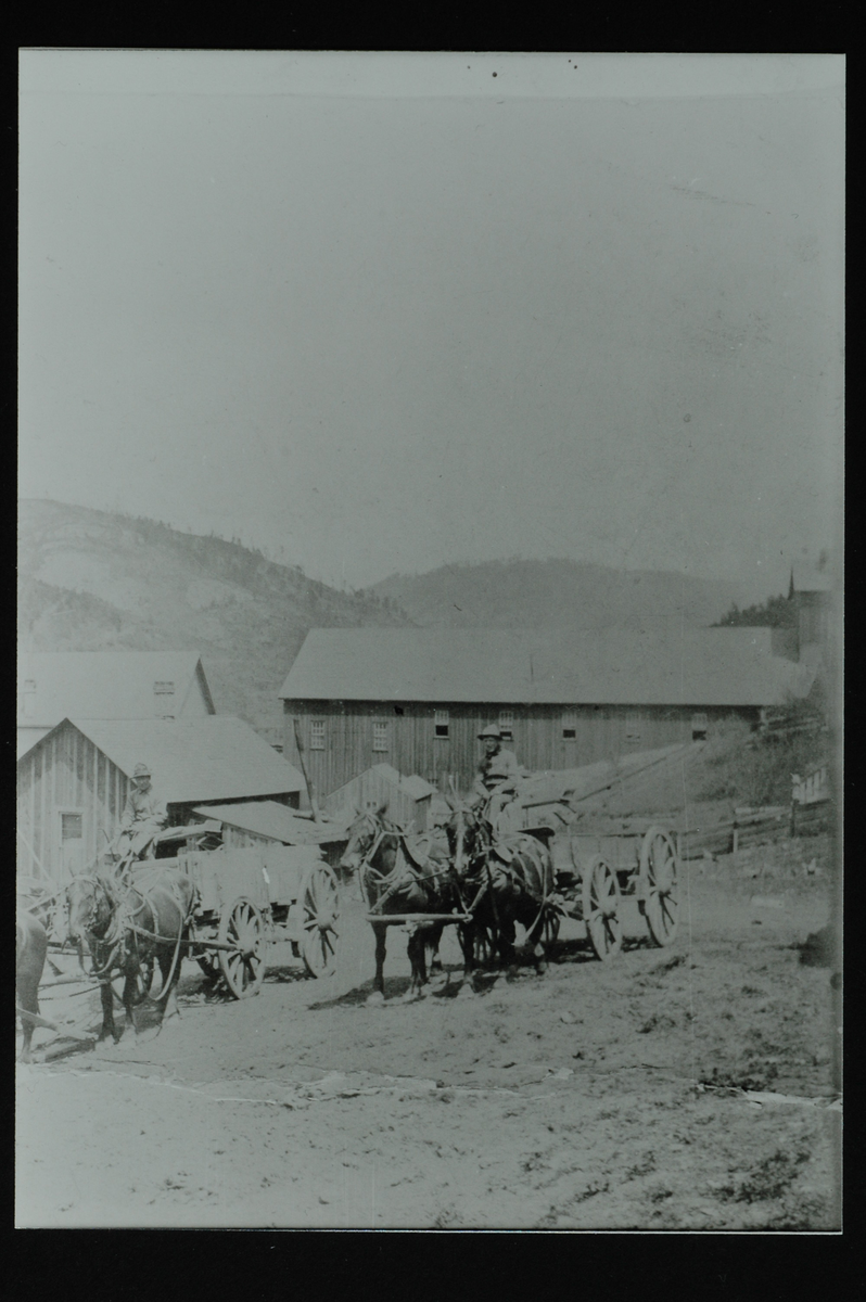 En gård med lys, to-etasjes hovedbygning, stor låve og flere uthus. I forgrunn to vogner med tilsammen fem hester foran. Til venstre en mann som leier en hest med et barn på. I bakgrunn en åsrygg med spredte trær. 