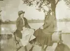 Två glada tonårsflickor i halvlånga kappor och hatt sitter uppflugna på en parksoffa vid Växjösjön, en solig vårdag. I bakgrunden skymtar man några byggnader på Öster.