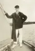 En ung man i seglarkostym med kritade vita skor, poserar i fören på en segelbåt.