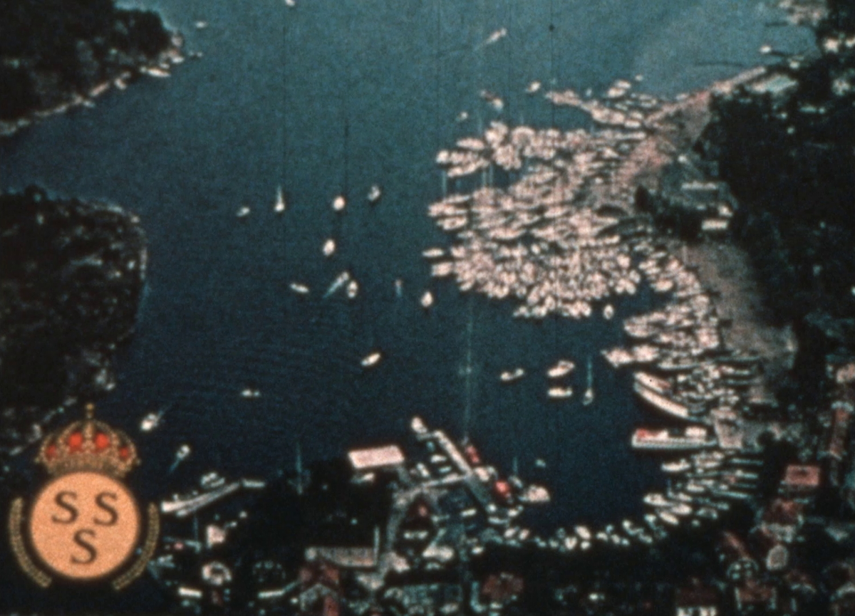 Färgfilm av Knud H. Reimers från Kungliga Svenska Segel Sällskapets (KSSS) jubileumsregatta i Sandhamn 1980, då KSSS firade 150 år. 


Segelbåten ASHANTI II ligger lastad på en vagn intill ett tågspår. Färden bär sedan av med båt mellan skärgårdsöar ut mot Sandhamn. Några av dem lägger man till vid längsmed vägen. Framme på Sandhamn vajar flera olika länders flaggor i vinden. Det hålls ett inledande tal på både tyska och svenska, varefter en representant för den internationella 30 kvm klassen av segelbåtar håller tal på engelska. Några av de närvarande är klädda i historisk stil. Arbete utförs i masten på en av segelbåtarna i hamnen. En segelbåt med norsk flagga utsirad med kung Olav V av Norges monogram ligger också förtöjd vid kajen. Flera tävlingar genomförs där ett stort antal båtar deltar. Mellan evenemangen bordar flera av båtarna varandra i en lång rad ute på vattnet. Från dem dyker och simmar besättningsmedlemmarna, minglar och bjuder varandra på alkohol över relingarna. I ytterligare kappseglingar har flera av de tävlande färgglada spinnakrar.


På land har en grupp personer gått ihop för att med gott humör lyfta och kasta någon annan från bryggan ner i vattnet mellan båtarna. Det har samlats en publik intill kajen och snart drar en prisceremoni igång. Vissa har videokameror med sig. Ett skott avfyras från en salutkanon och tal hålls. Sedan är det dags för avfärd från Sandhamn. Man tar med sig packning och vinkar farväl i hamnen innan båtarna ger sig av mot vad som skulle kunna vara Kanholmsfjärden, där regattans slutkläm med samtliga båtar närvarande är planerad att gå av stapeln.