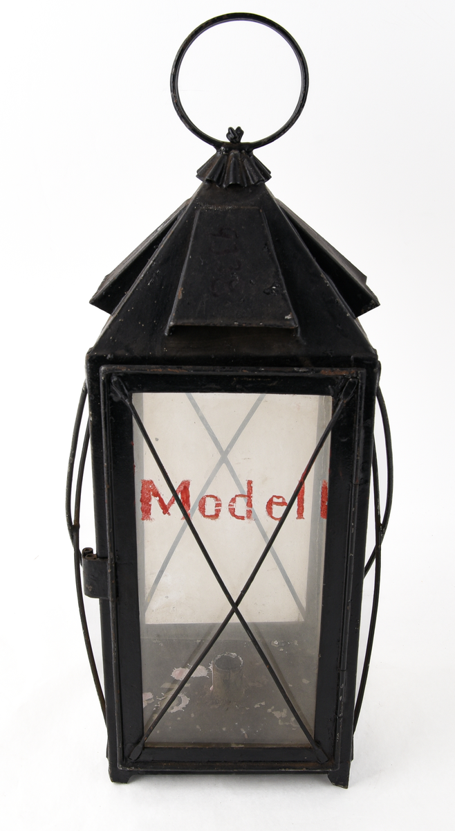 Rektangulär truppvagnslykta av svart metall. Stor ögla och ornamenterat rökutsläpp upptill. Fyra, rektangulära, klara glasskivor. En av glasen är märkt med text i rött "Modell" och fäst på en öppningsbar lucka för att komma åt hållaren för stearinljus. Staerinrester i botten av lyktan. Tunna, korslagda metalltrådar framför glasskivorna.