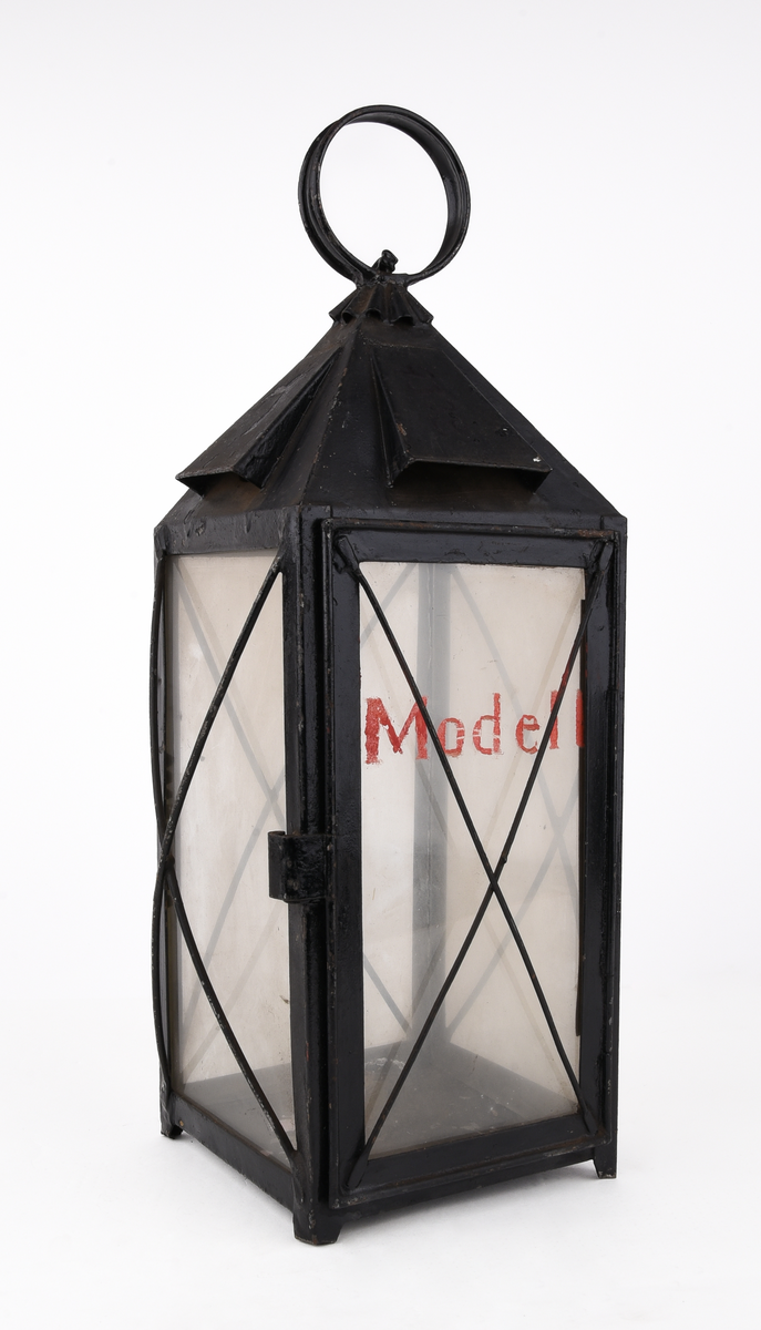 Rektangulär truppvagnslykta av svart metall. Stor ögla och ornamenterat rökutsläpp upptill. Fyra, rektangulära, klara glasskivor. En av glasen är märkt med text i rött "Modell" och fäst på en öppningsbar lucka för att komma åt hållaren för stearinljus. Staerinrester i botten av lyktan. Tunna, korslagda metalltrådar framför glasskivorna.