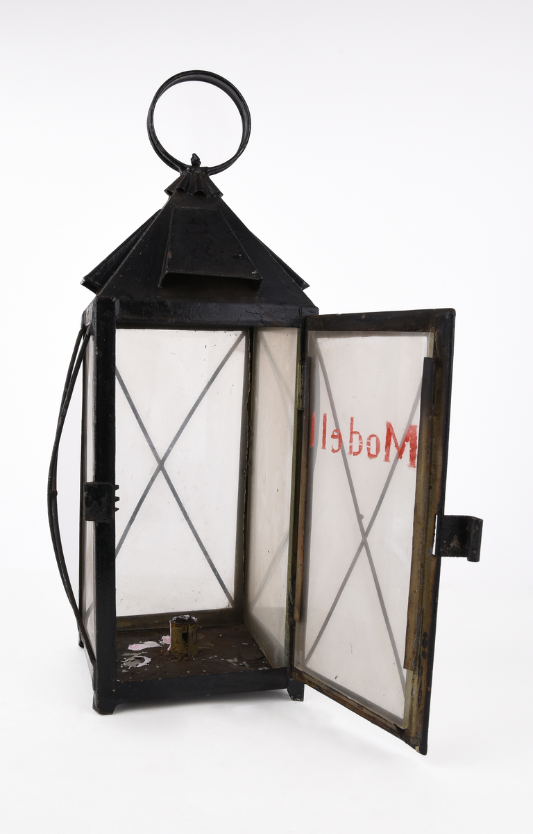 Rektangulär truppvagnslykta av svart metall. Stor ögla och ornamenterat rökutsläpp upptill. Fyra, rektangulära, klara glasskivor. En av glasen är märkt med text i rött "Modell" och fäst på en öppningsbar lucka för att komma åt hållaren för stearinljus. Staerinrester i botten av lyktan. Tunna, korslagda metalltrådar framför glasskivorna.
