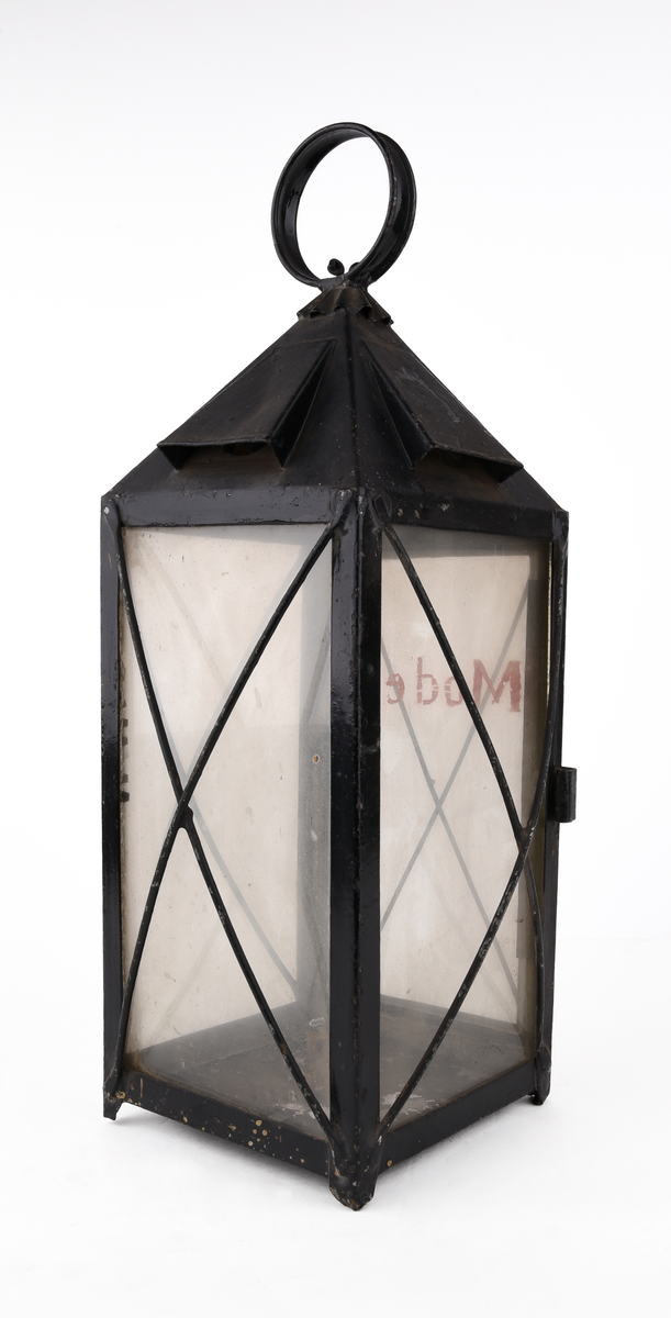 Rektangulär truppvagnslykta av svart metall. Stor ögla och ornamenterat rökutsläpp upptill. Fyra, rektangulära, klara glasskivor. En av glasen är märkt med text i rött "Modell" och fäst på en öppningsbar lucka för att komma åt hållaren för stearinljus. Staerinrester i botten av lyktan. Tunna, korslagda metalltrådar framför glasskivorna.