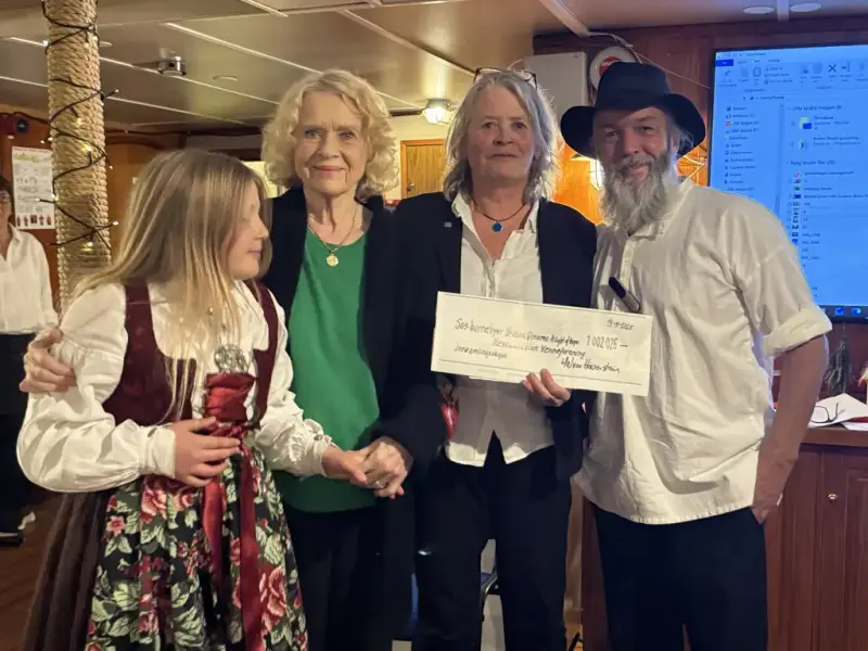 En kvinne holder opp en sjekk. Ei jente i festdrakt og en kvinne i grønn overdel står til venstre for henne, til høyre en mann med hatt og skjegg. Et skipsinteriør i bakgrunn.