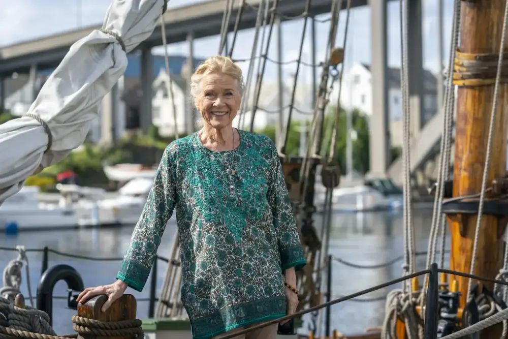 En kvinne i mønstret grønn overdel står ombord en seilskute. En bru og bygninger i bakgrunn. 