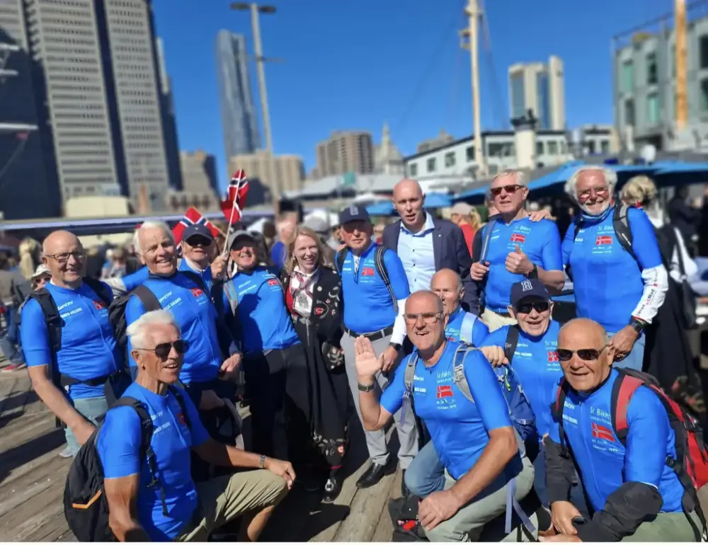 En gruppe menn i blå overdeler og en dame i bunad i midten. To små norske flagg holdes opp. Høyhus i bakgrunn. 