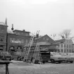 Nedslag på Stora torget i Linköping, sannolikt 1970. Kvartet Apotekets hus i fonden har ansetts tjänat ut och längans köpmän realiserar inför kommande rivning.