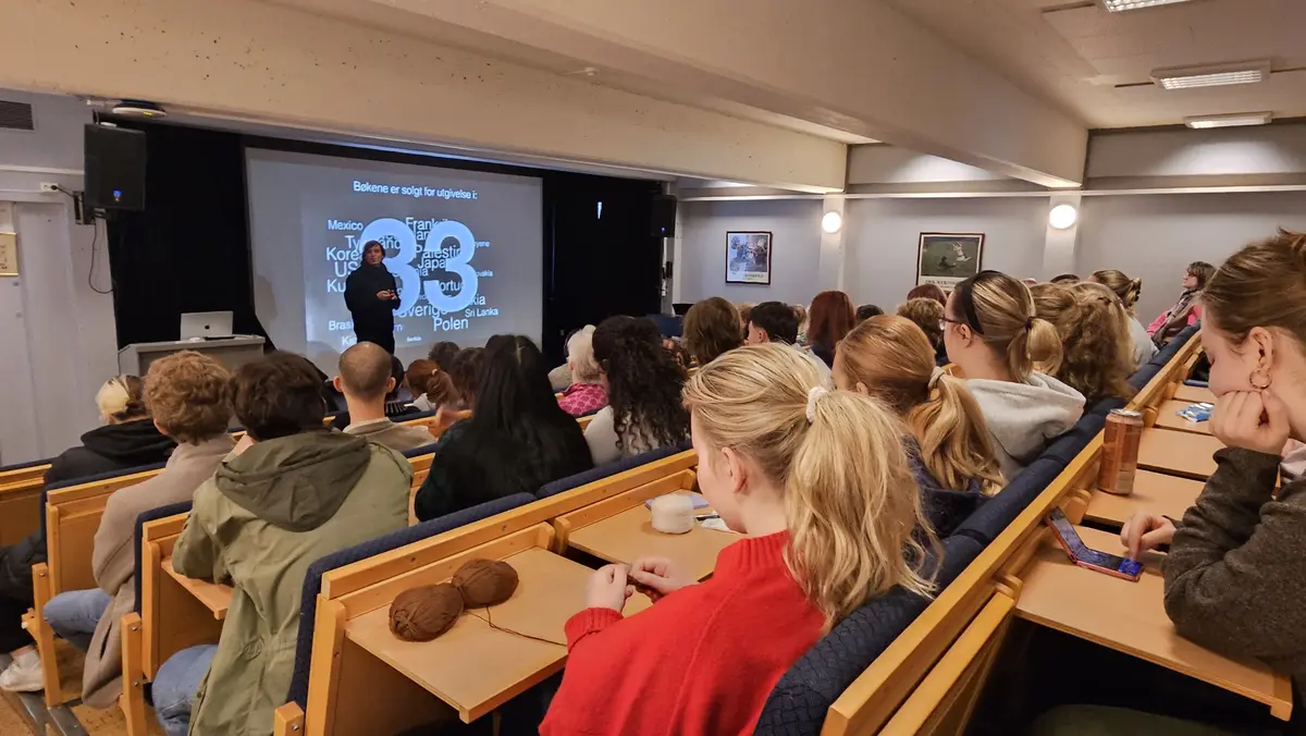 Unge mennesker sitter i et auditorium og ser mot en foredragsholder foran en skjerm til venstre.