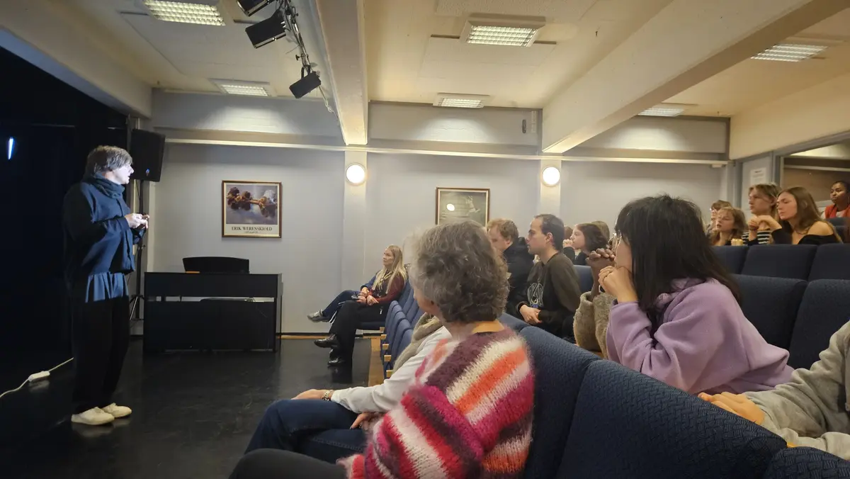 En mann står til venstre i et auditorium og snakker mot publikum som sitter til høyre. 